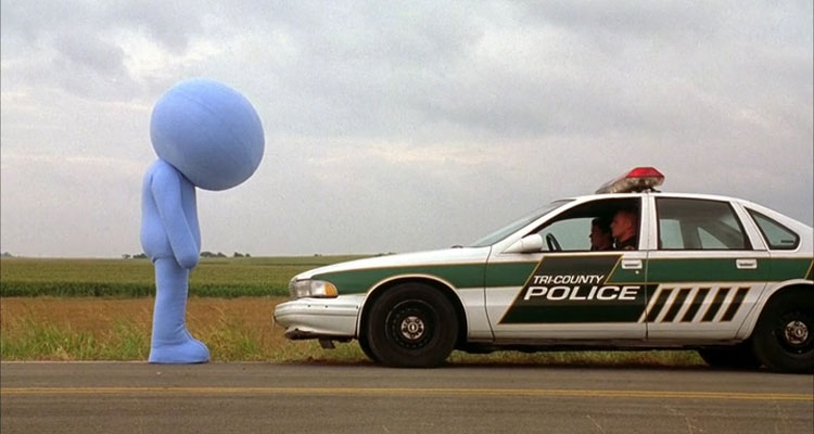 Kabluey 2007 Movie Scene Scott Prendergast as Salman dressed in a giant blue suit standing in front of a police car