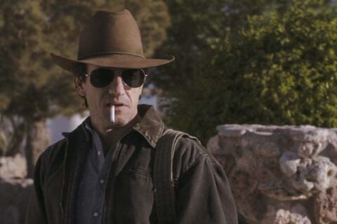 The Rambler 2013 Movie Scene Dermot Mulroney as The Rambler, a drifter who just got out of prison, smoking a cigarette and wearing a cowboy hat and sunglasses