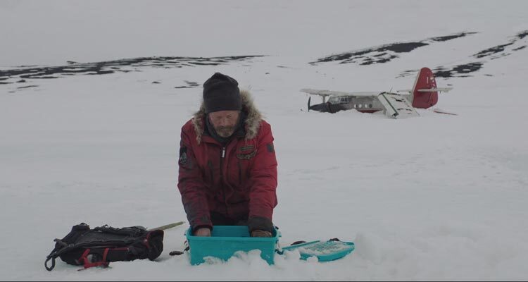 Arctic 2018 Movie Scene Mads Mikkelsen as Overgård stacking the fish he caught next to the wreckage of his plane