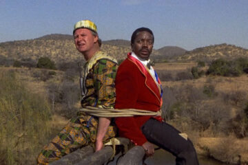 Yankee Zulu 1993 Movie Leon Schuster as Rhino and John Matshikiza as Zulu sitting on a plank with an elephant on the other side
