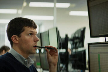 The Hummingbird Project 2018 Movie Scene Jesse Eisenberg as Vincent Zaleski holding a pencil in his mouth and looking at the monitors