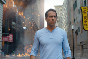 Free Guy 2021 Movie Scene Ryan Reynolds as Guy wearing a blue shirt with a building exploding behind him