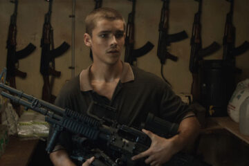Son of a Gun 2014 Movie Scene Brenton Thwaites as JR holding a heavy machinegun with AK-47 behind him