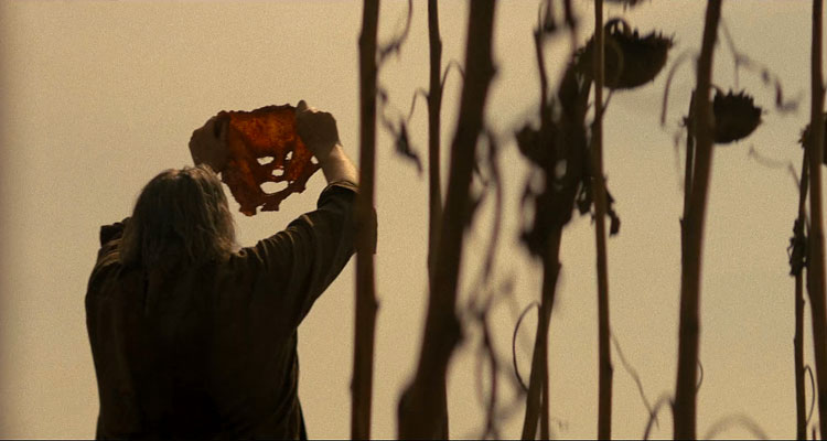 Texas Chainsaw Massacre 2022 Movie Scene Mark Burnham as Leatherface holding a mask made out of human skin above his head in a field of sunflowers