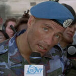 Street Fighter 1994 Movie Scene Jean-Claude Van Damme as Colonel Guile holding a mic and talking to the screen while wearing camo and a blue beret