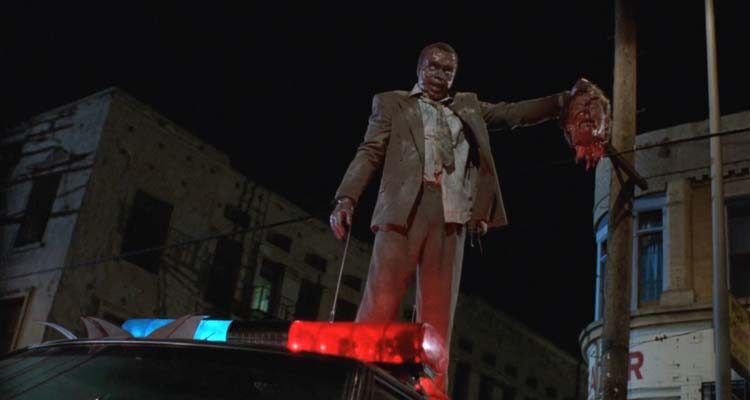 Tales From The Hood 1995 Movie Scene Tom Wright as Zombie Martin Moorehouse holding the severed head of Wings Hauser as Strom and standing on top of a police car