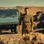 The Wages of Fear 2024 Movie Scene Alban Lenoir as Alex standing in front of a big rock with the truck carrying nitroglycerine in the background